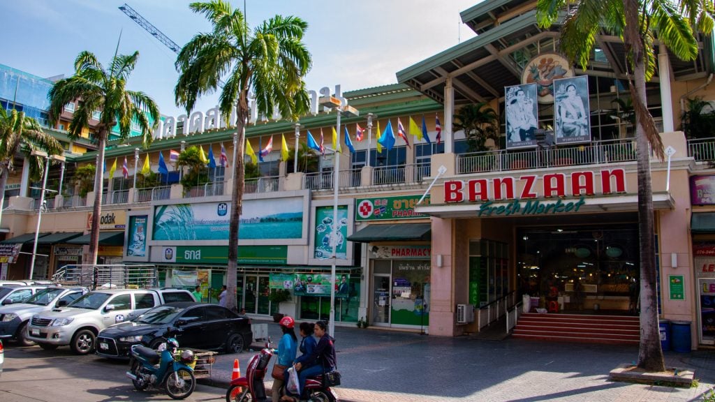 Banzaan Fresh Market Phuket exterior with entrance signage, flags, palm trees, parked cars, and motorbikes.