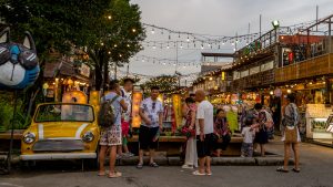 Chillva Market Phuket entrance with a parked car, people gathering, two-level market buildings, and hanging string lights.