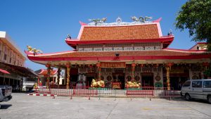Front view of Am Bang Neow Shrine with a red-tiled roof, dragon figures along the ridge, hanging red lanterns, and a wide concrete forecourt.