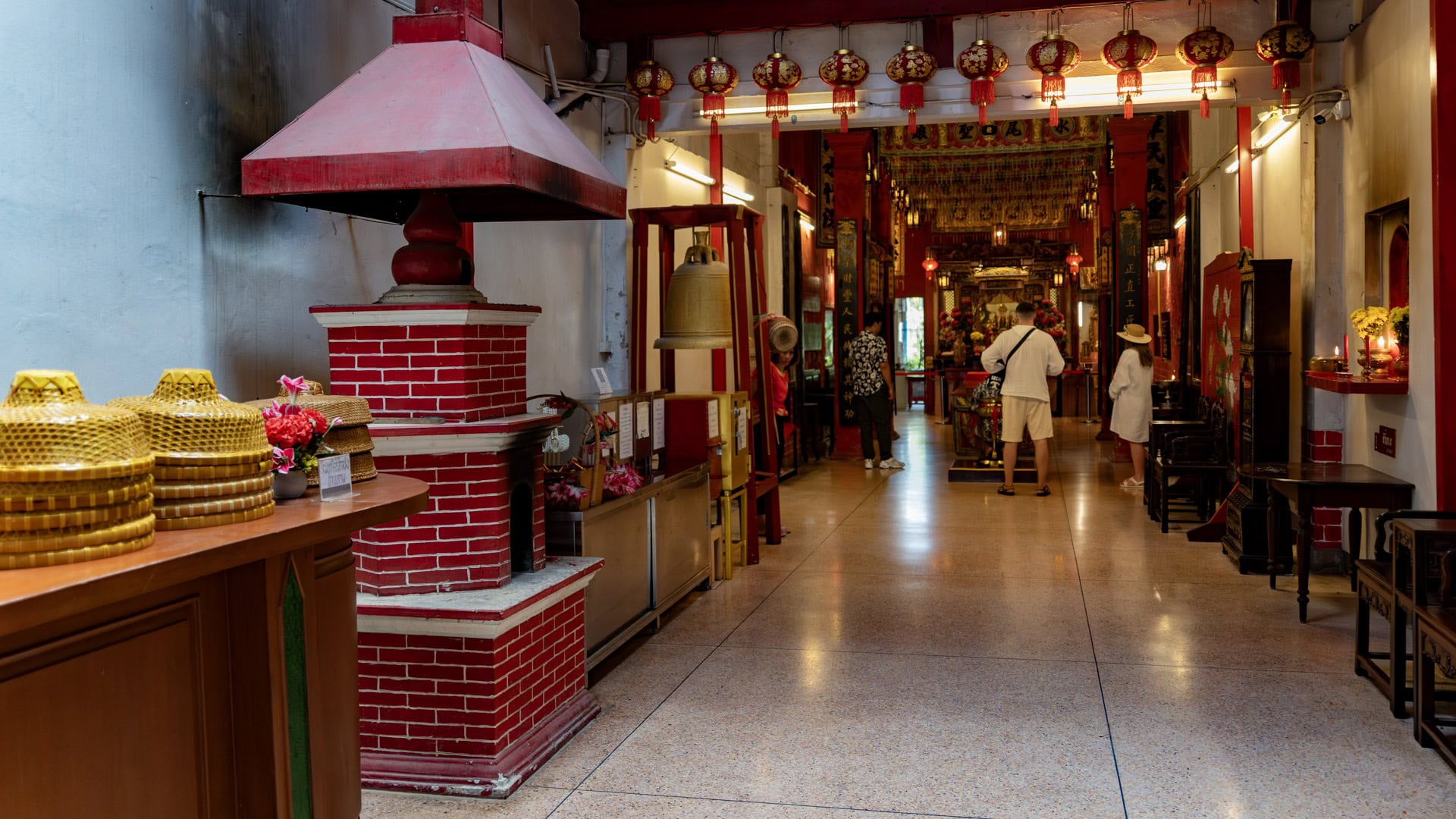 A long interior hall leads toward the shrine altar, with a large bell on a wooden frame, hanging lanterns, and a red brick incense chimney on the left.