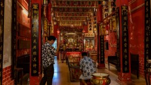 Two men stand and kneel before the main altar inside the shrine, surrounded by red pillars, gold calligraphy panels, and hanging banners.