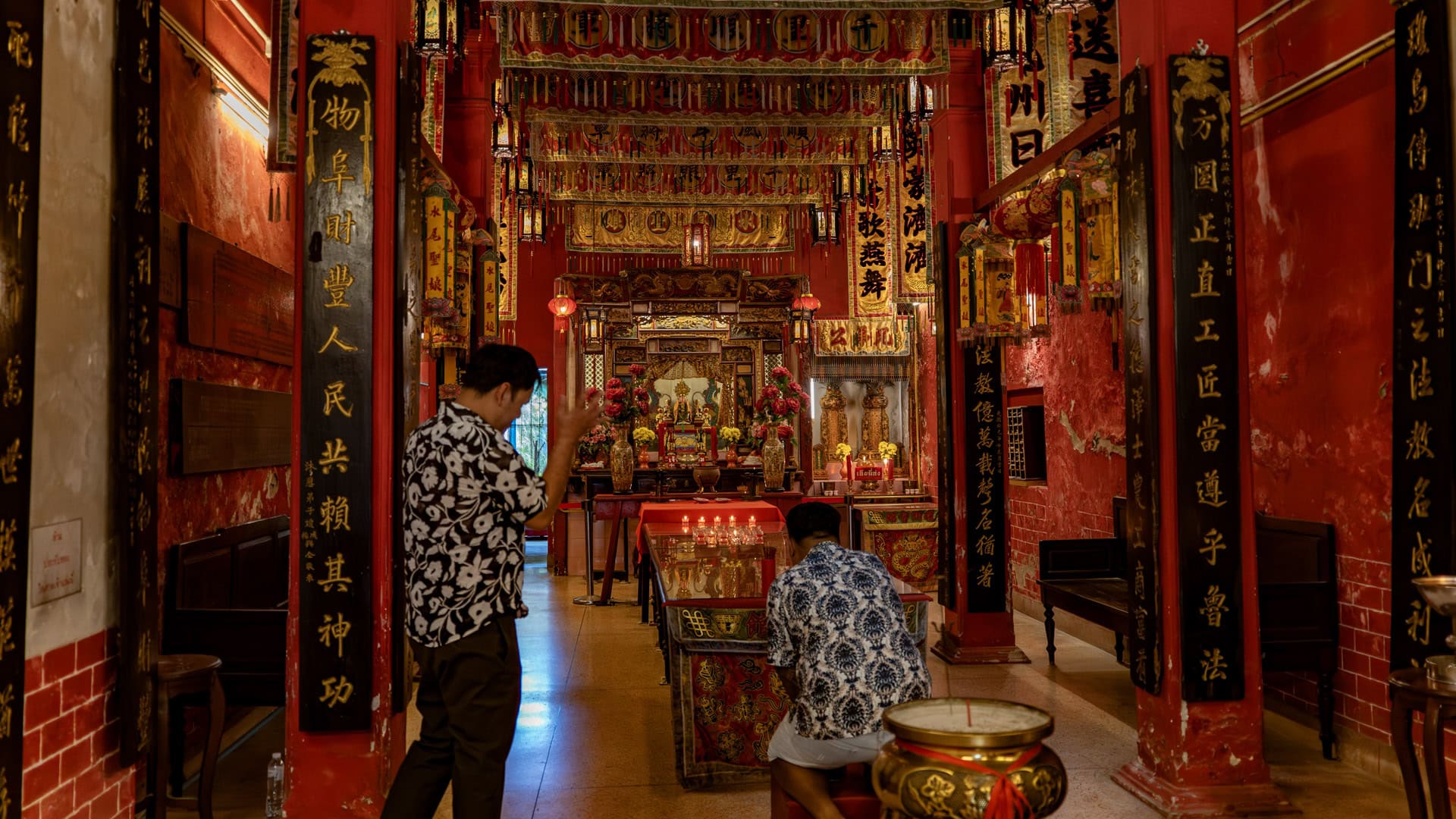 Two men stand and kneel before the main altar inside the shrine, surrounded by red pillars, gold calligraphy panels, and hanging banners.