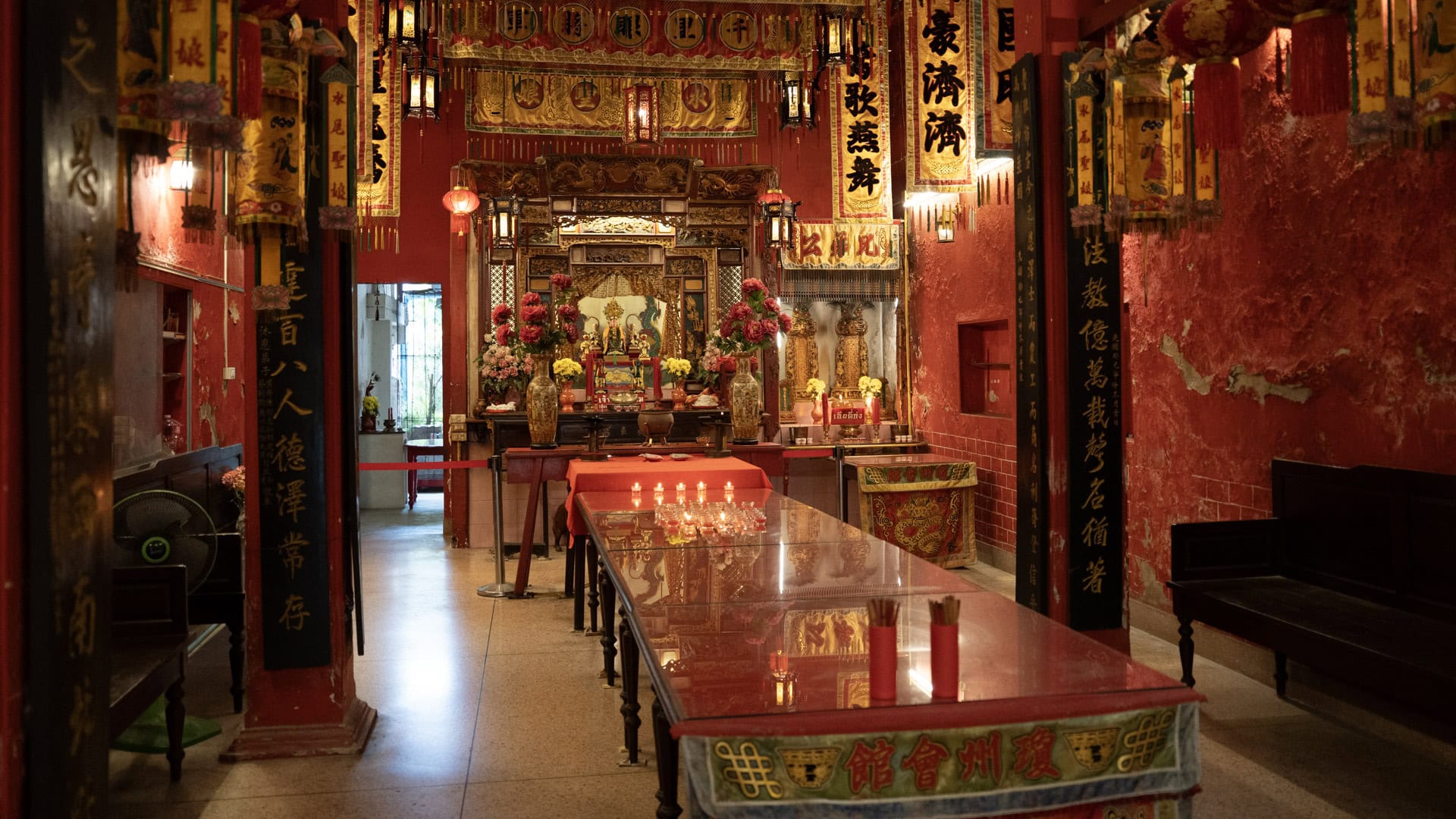 A red-topped offering table stands in the centre of the shrine hall, leading toward the main altar between red walls, calligraphy pillars, and hanging banners.