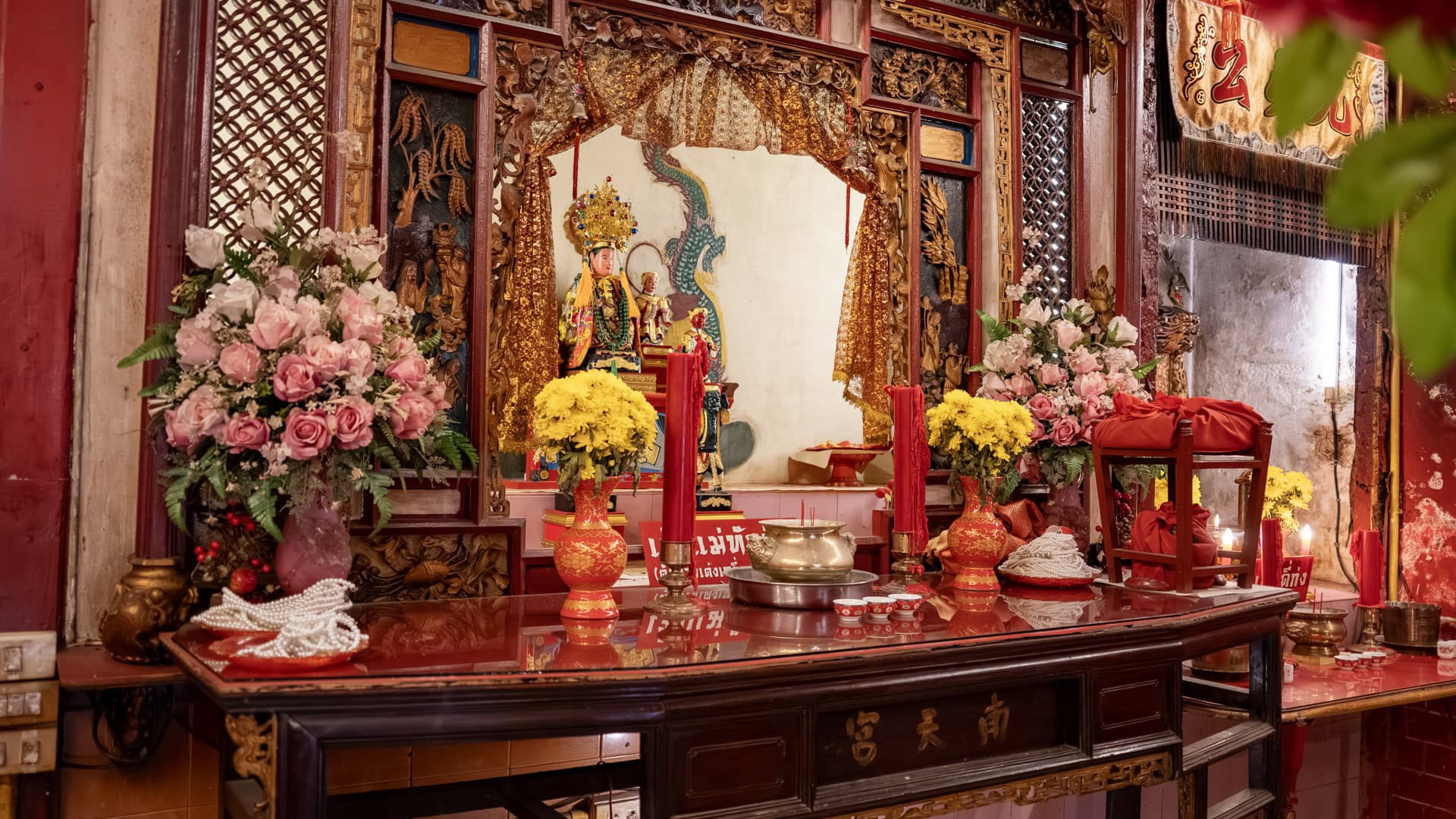 A side altar displays a seated deity figure behind carved woodwork, flanked by pink roses, yellow chrysanthemums, red candles, and offering vessels.