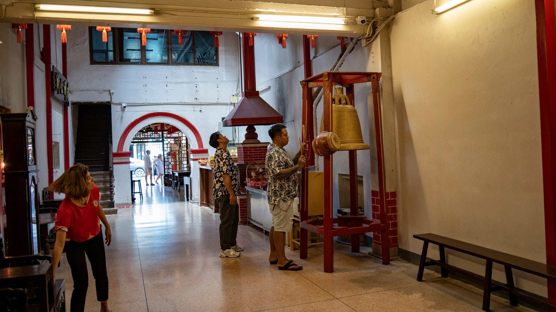 A man strikes a hanging bell in the shrine’s front hall while two other people stand nearby beneath red beams and lanterns.