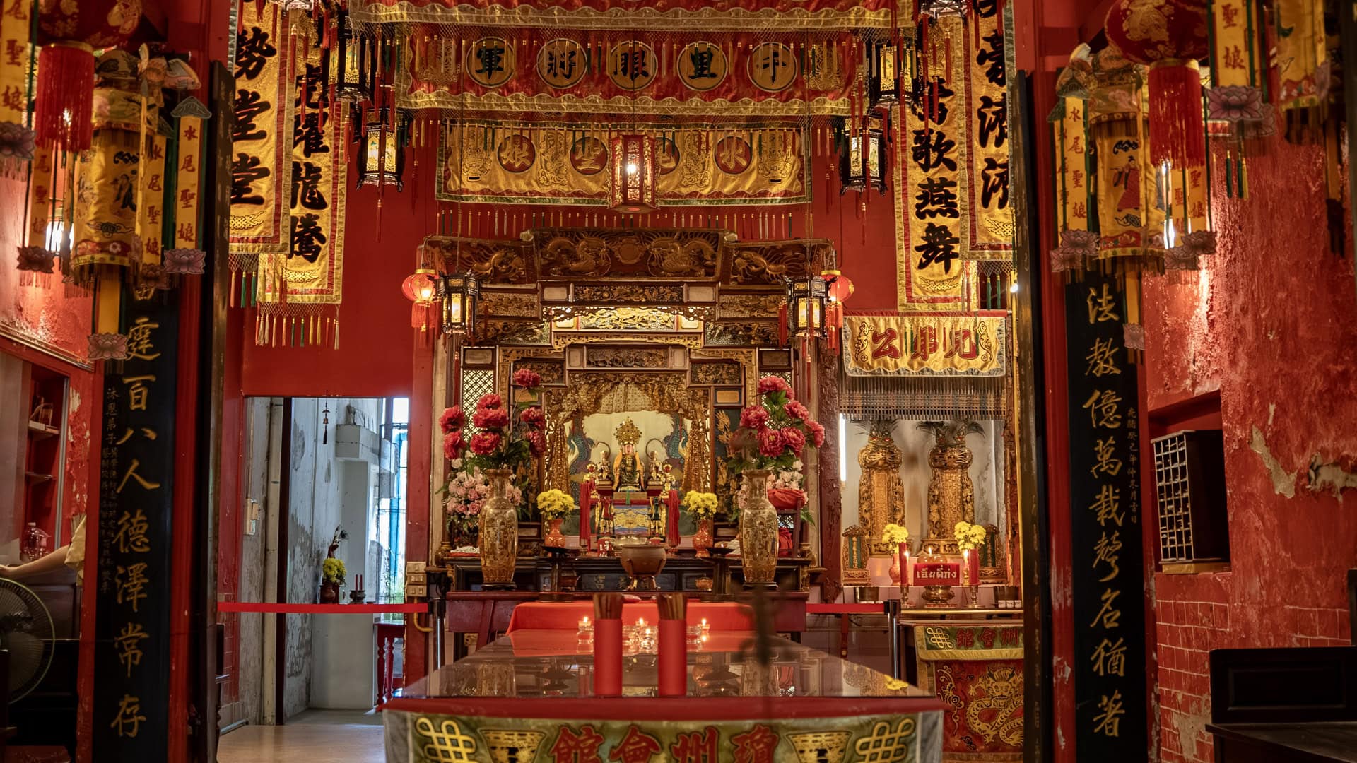 The main altar stands beneath layered hanging banners and lanterns, with flower vases, candles, and black calligraphy pillars framing the shrine hall.
