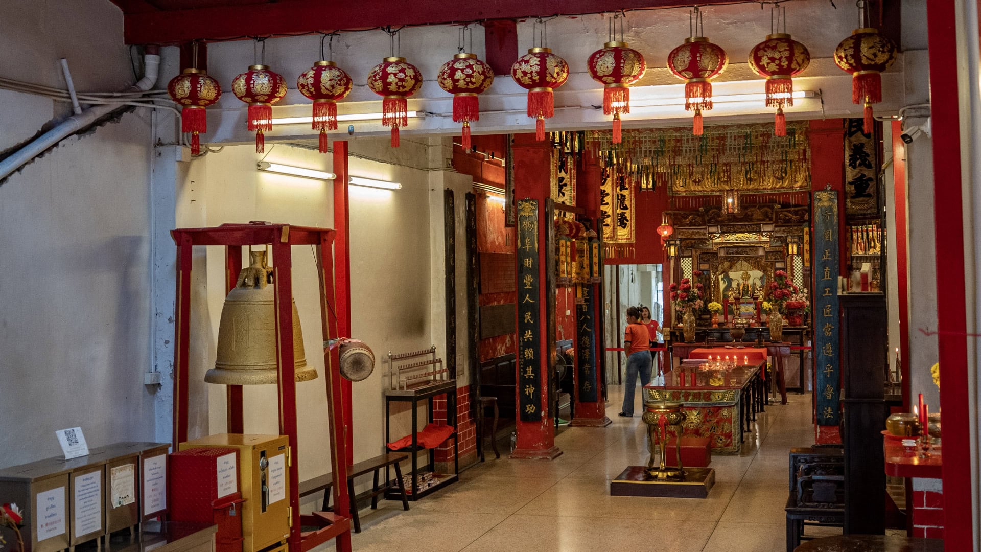 A side view of the shrine hall shows a hanging bell on the left, a row of red lanterns overhead, and the main altar visible in the distance.