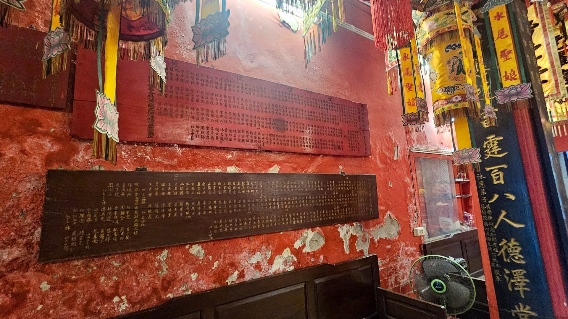 Dark wooden inscription panels hang on a worn red wall beneath suspended banners, beside a black calligraphy pillar and a floor fan.