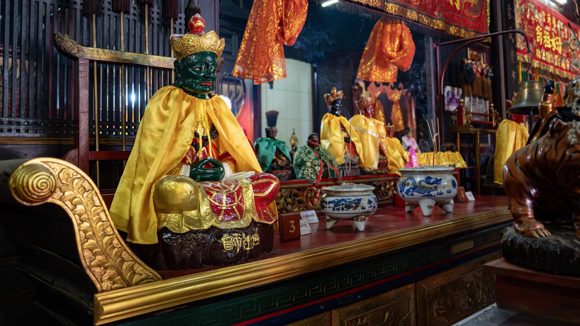 Several seated shrine figures in yellow robes line a wooden altar inside the shrine, with incense burners, red hanging cloths, and smoke visible above them.