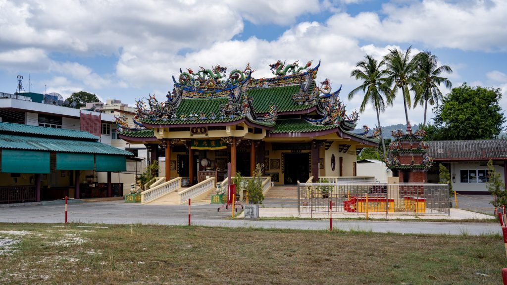 A Chinese shrine with green tiled roofs, dense dragon roof ornament, and palm trees behind the building.
