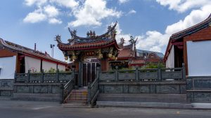 The front shrine hall at San Chao Jui Tui stands behind a grey stone fence with carved relief panels, with curled roof ridges and small deity figures along the roofline.
