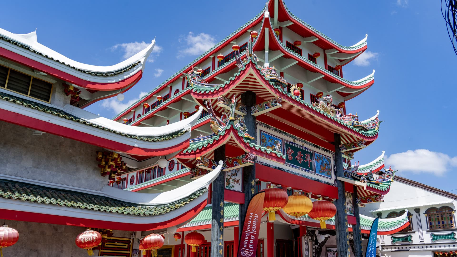At San Chao Kathu, an ornate red shrine gate with hanging lanterns stands in front of a multi-storey building with curved green-tiled rooflines and decorative roof ornaments.