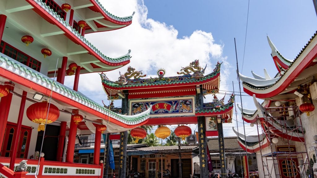At San Chao Kathu, an ornate inner gate with dragons on the roof crest spans the space between shrine buildings, with red and yellow lanterns hanging underneath.