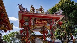 The entrance arch roof at San Chao Kiew Tien Keng carries twin dragon ornaments and hanging red lanterns, with a large tree behind it.