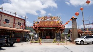 Front view of Sam Kong Shrine with an open forecourt, red lantern strings, dragon roof ornaments, black plaques, and parked vehicles on both sides.
