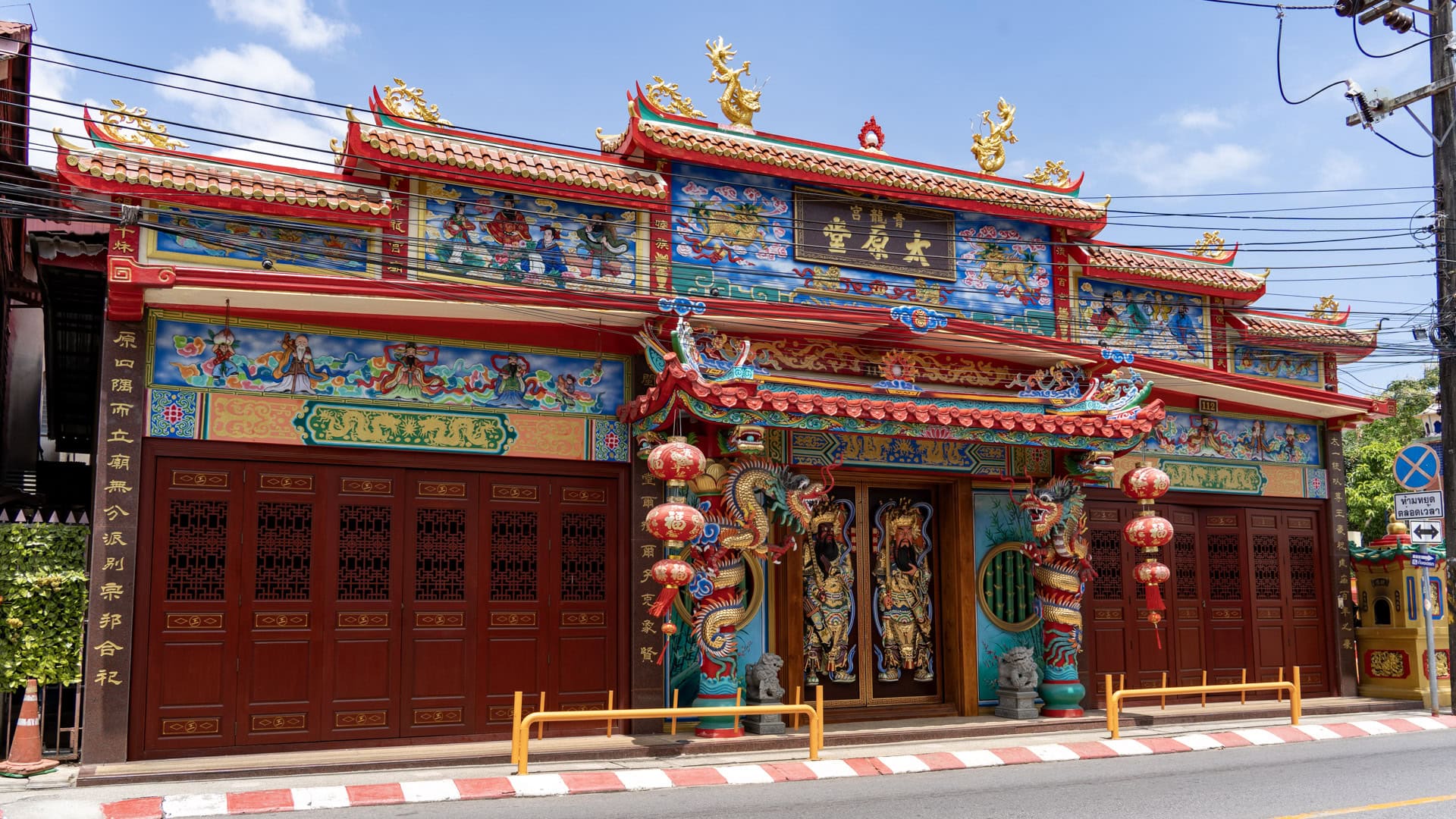 The street-facing façade of San Chao Mae Yanang shows a red entrance porch, painted mythological panels, dragon-wrapped columns, and rows of wooden doors beneath overhead wires.