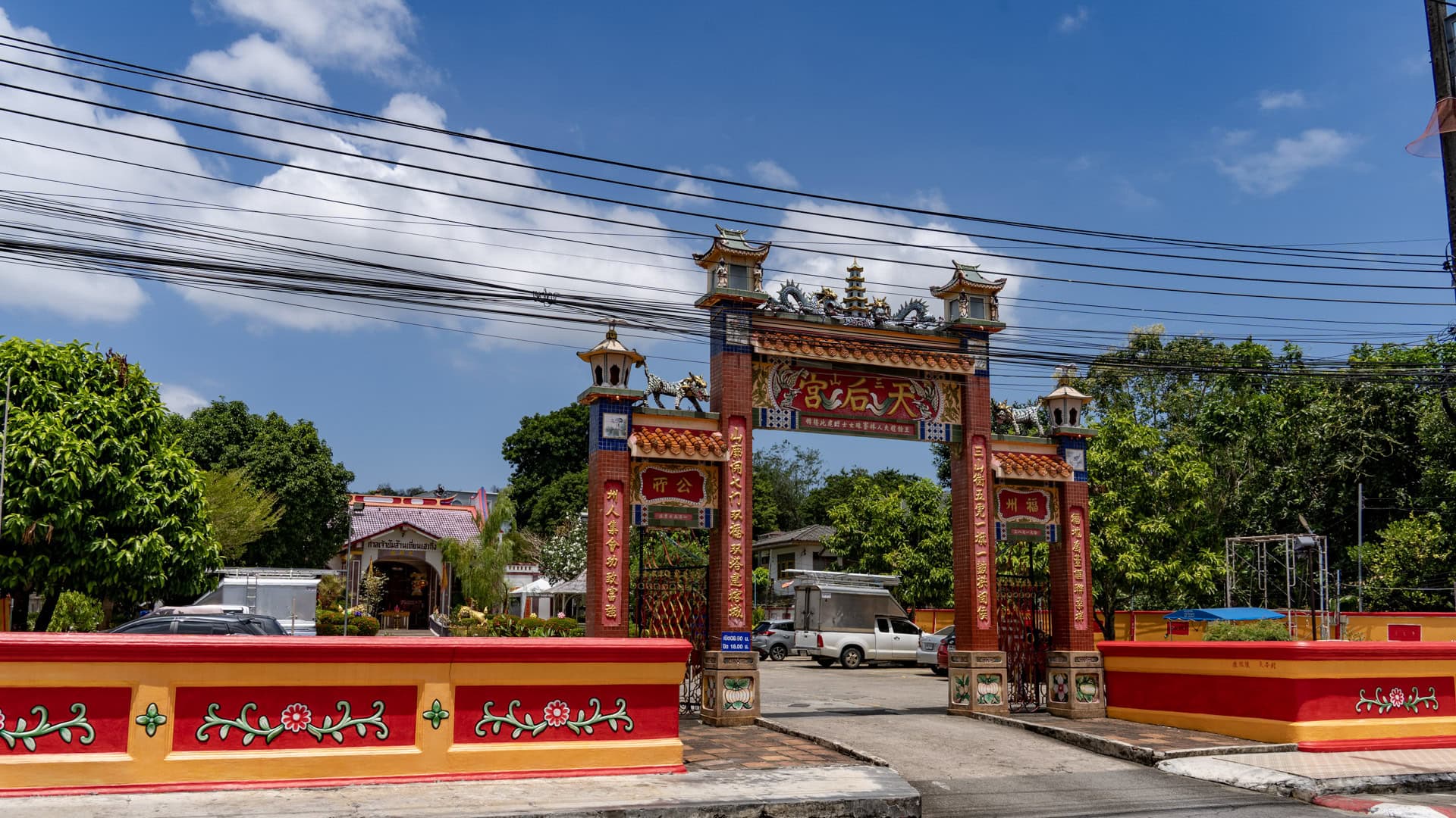 The entrance gate at San Chao Mae Yanang stands behind a red-and-yellow boundary wall, with Chinese characters on the pillars, tiled roof details, and parked vehicles inside the compound.