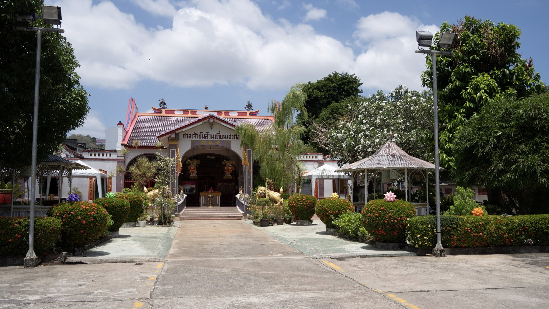 The main hall at San Chao Mae Yanang stands at the end of a concrete courtyard, with clipped flowering shrubs, gold guardian figures, and a covered porch below a tiled roof.