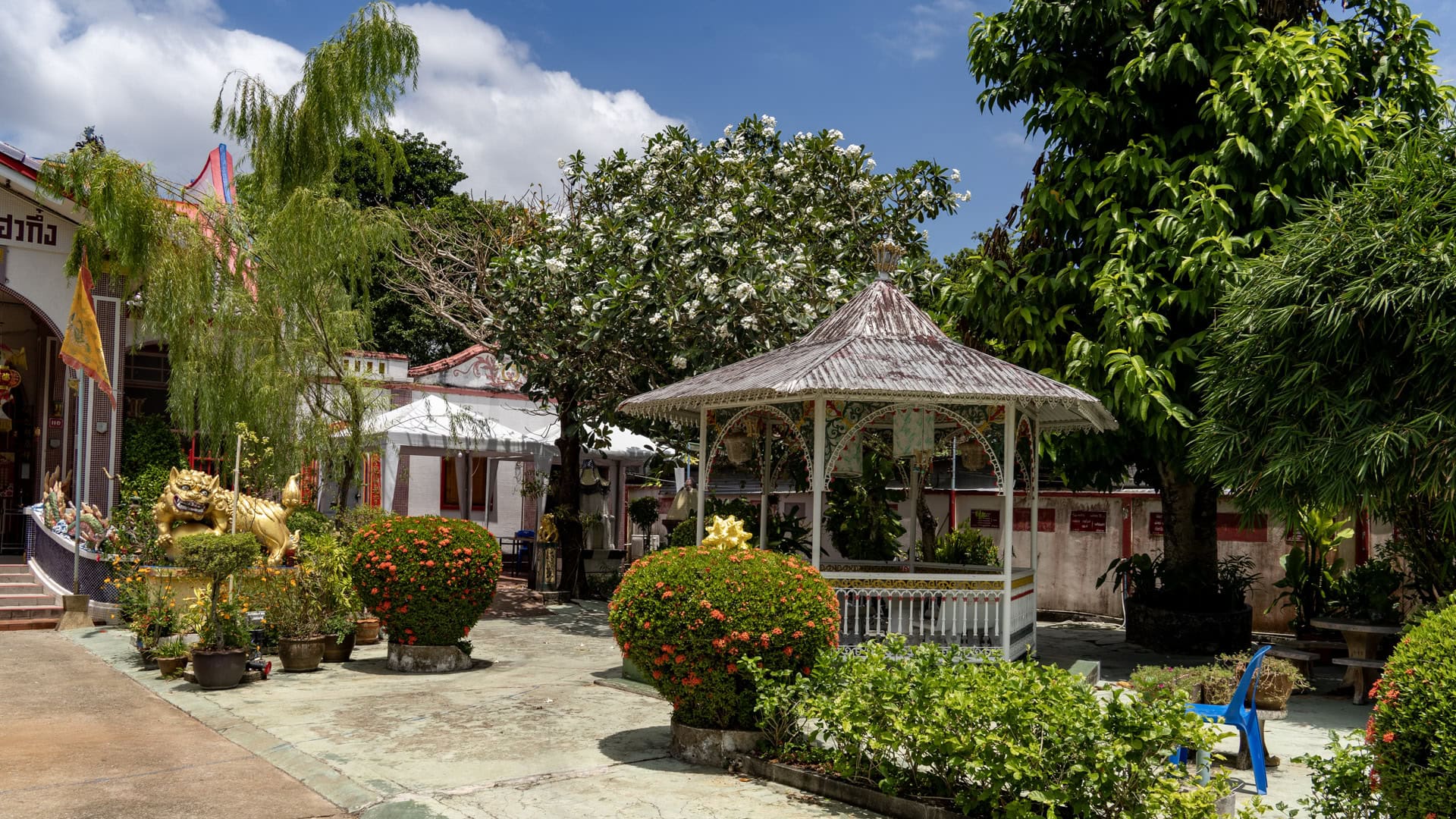 A white metal pavilion at San Chao Mae Yanang sits among flowering shrubs and tall trees, with part of the shrine hall and a gold guardian figure visible to the left.
