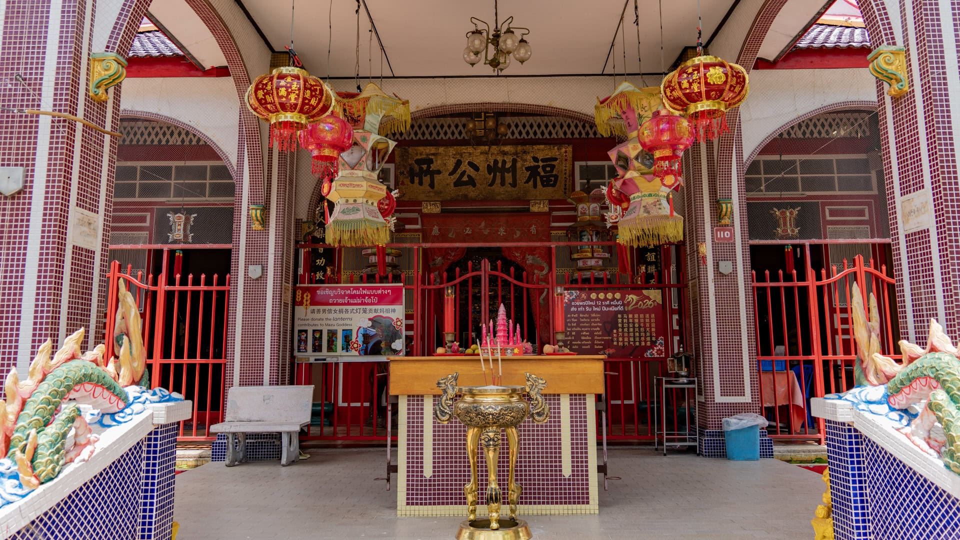 The entrance porch at San Chao Mae Yanang has red lanterns, hanging festival lamps, a brass incense urn, and red gates behind a wooden offering table.