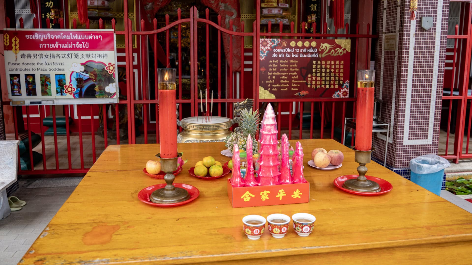 An offering table at San Chao Mae Yanang holds red candles, pink wax offerings, fruit plates, and tea cups, with red railings and shrine notices behind.