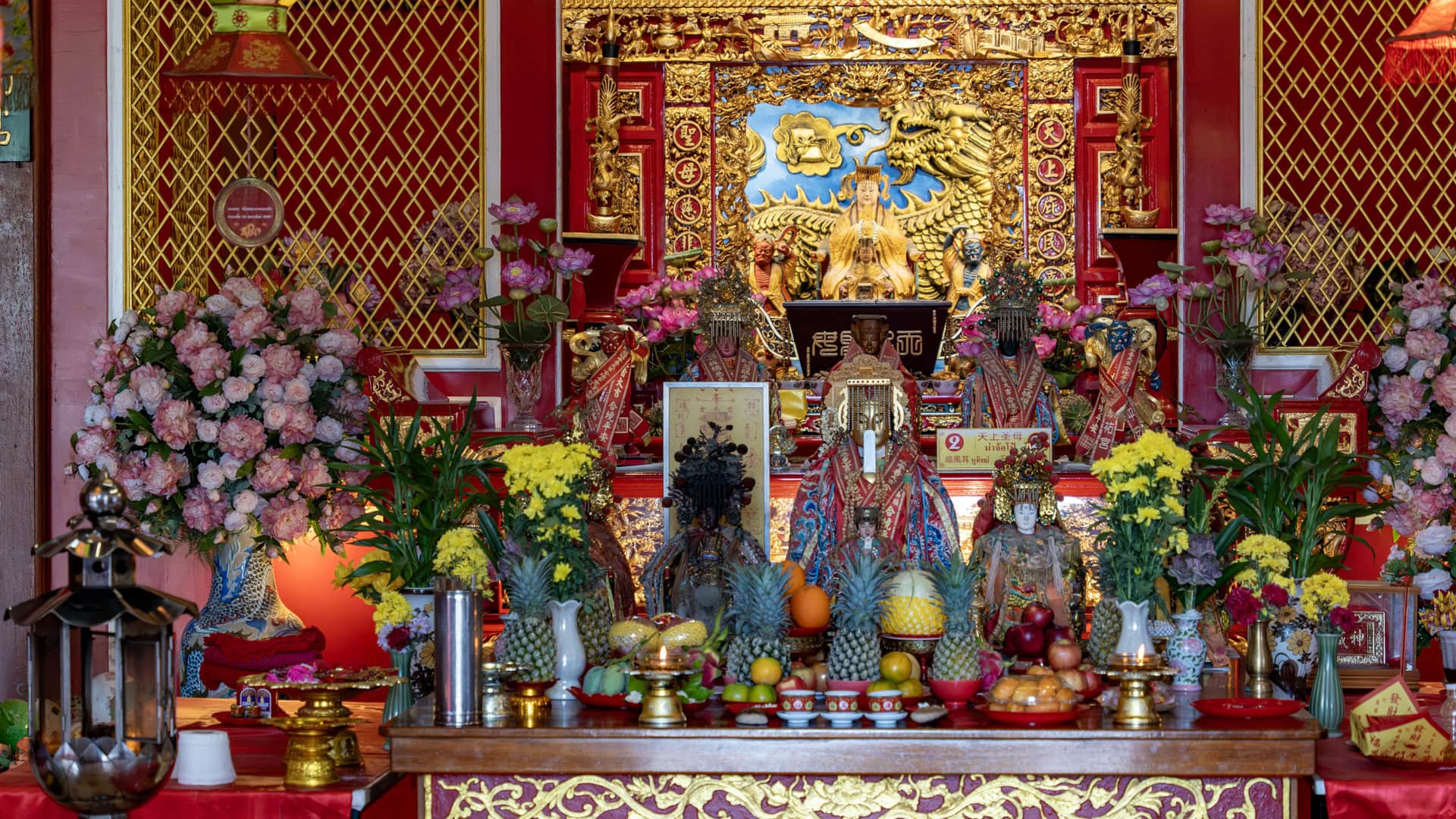 The main altar at San Chao Mae Yanang contains seated deity figures, pink and yellow flower arrangements, pineapples, and trays of fruit before a red-and-gold backdrop.