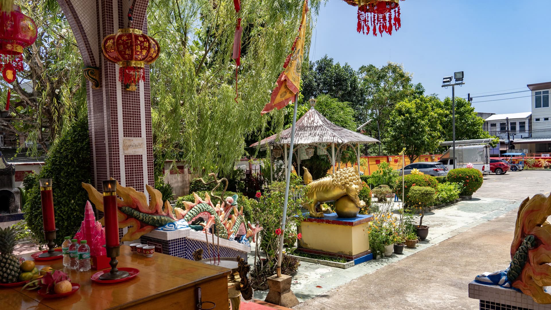 A side view from the porch at San Chao Mae Yanang shows dragon-edged rails, a gold guardian figure, a flag, a small pavilion, and planted shrubs beside the forecourt.
