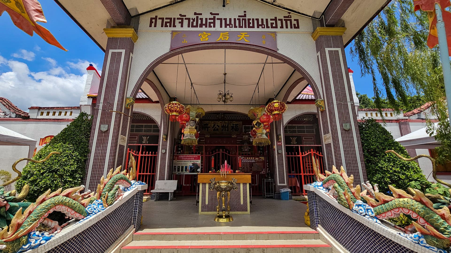 The front entrance of San Chao Mae Yanang is framed by a tall portico, red gates, hanging lanterns, and low dragon-shaped balustrades leading to an incense table.