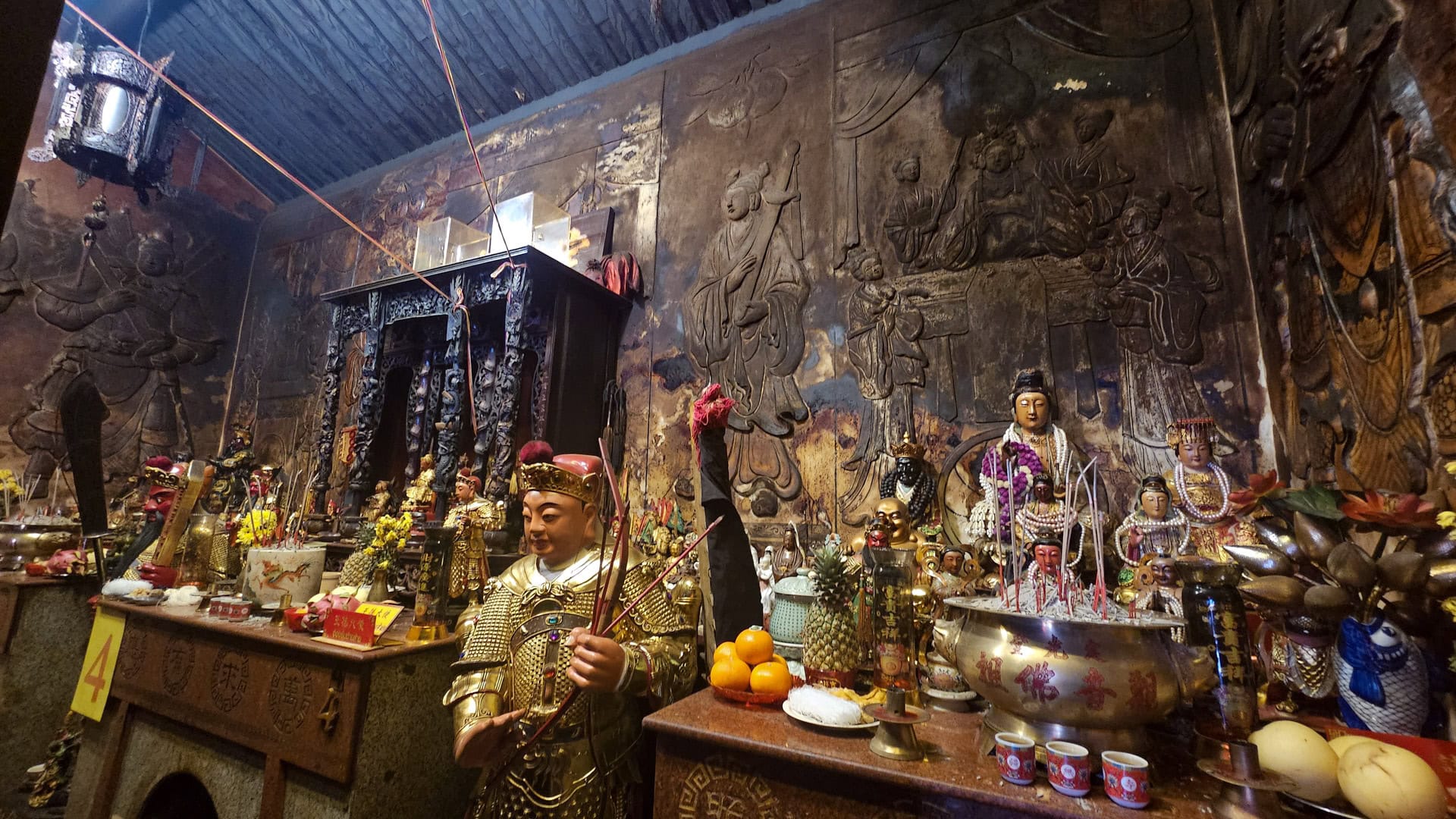 A guardian figure stands before a side altar at San Chao Saengtham, with incense bowls, fruit offerings, small statues, and large carved wall reliefs behind.