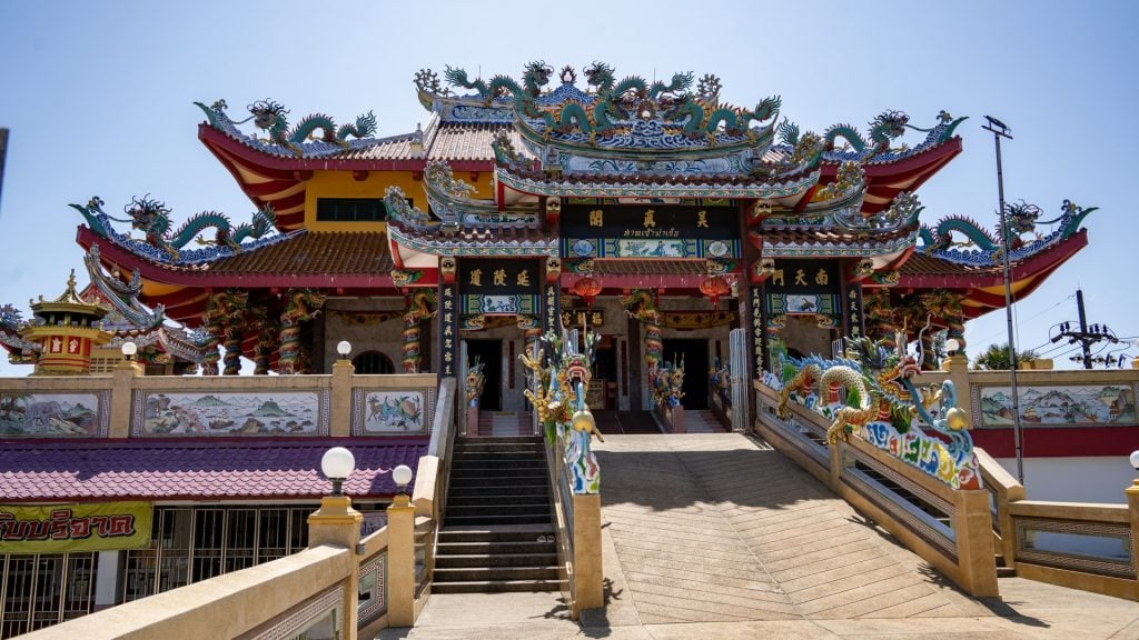 The main entrance of San Chao Tha Ruea is framed by layered rooflines with dragon ornaments, black signboards, and twin stairways rising to the hall.
