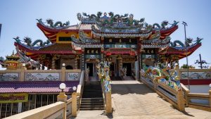 The main entrance of San Chao Tha Ruea is framed by layered rooflines with dragon ornaments, black signboards, and twin stairways rising to the hall.