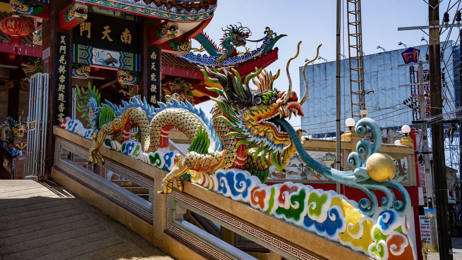 A long painted dragon runs along the balustrade of the entrance ramp at San Chao Tha Ruea, with shrine columns and roadside wires behind it.