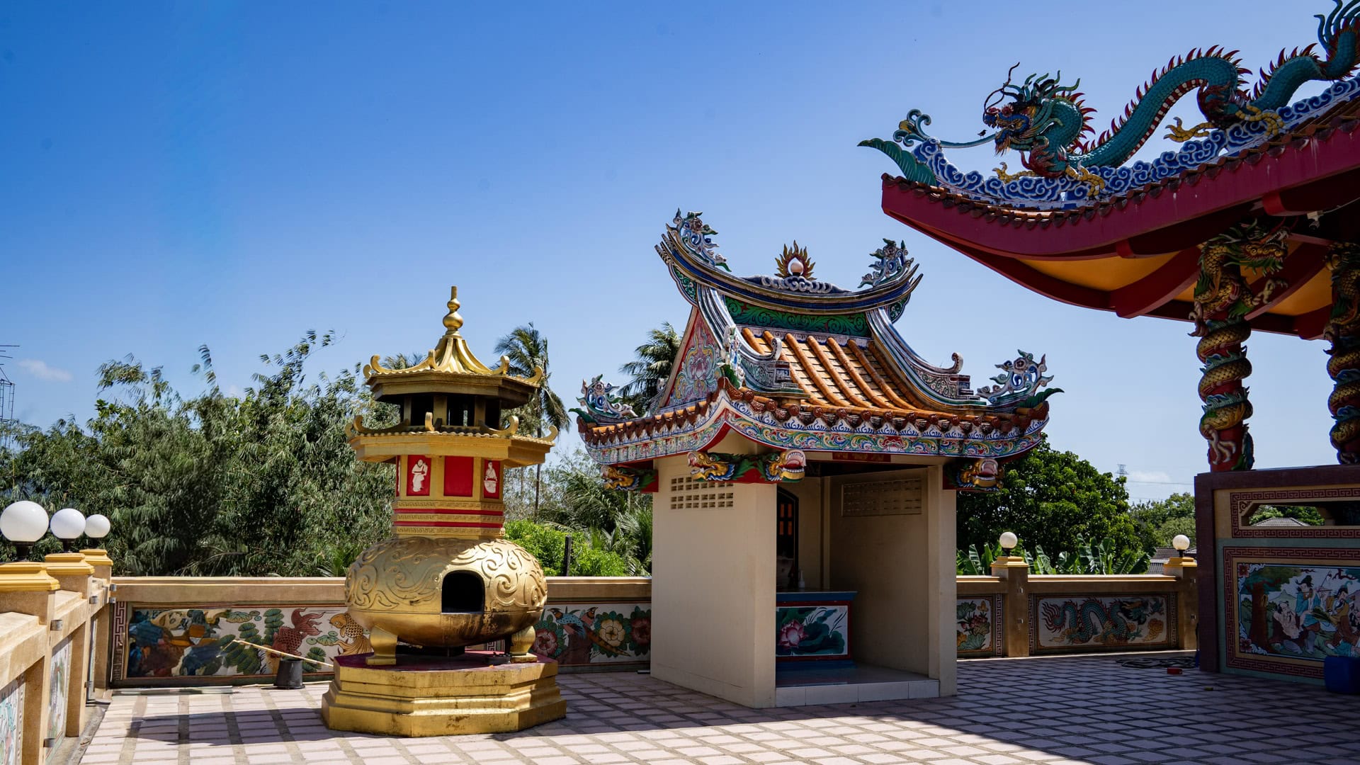 A gold incense burner stands beside a small side shrine at San Chao Tha Ruea, with tiled paving, painted roof trim, and trees beyond the courtyard wall.