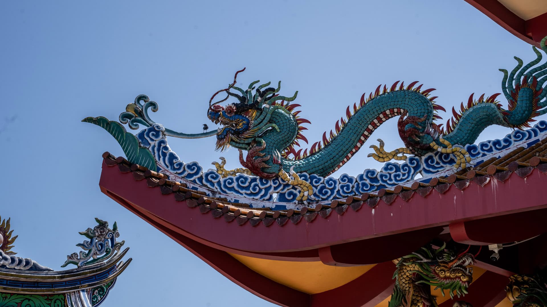 A turquoise dragon ornament crouches along the red roof edge at San Chao Tha Ruea, surrounded by blue cloud motifs against a clear sky.