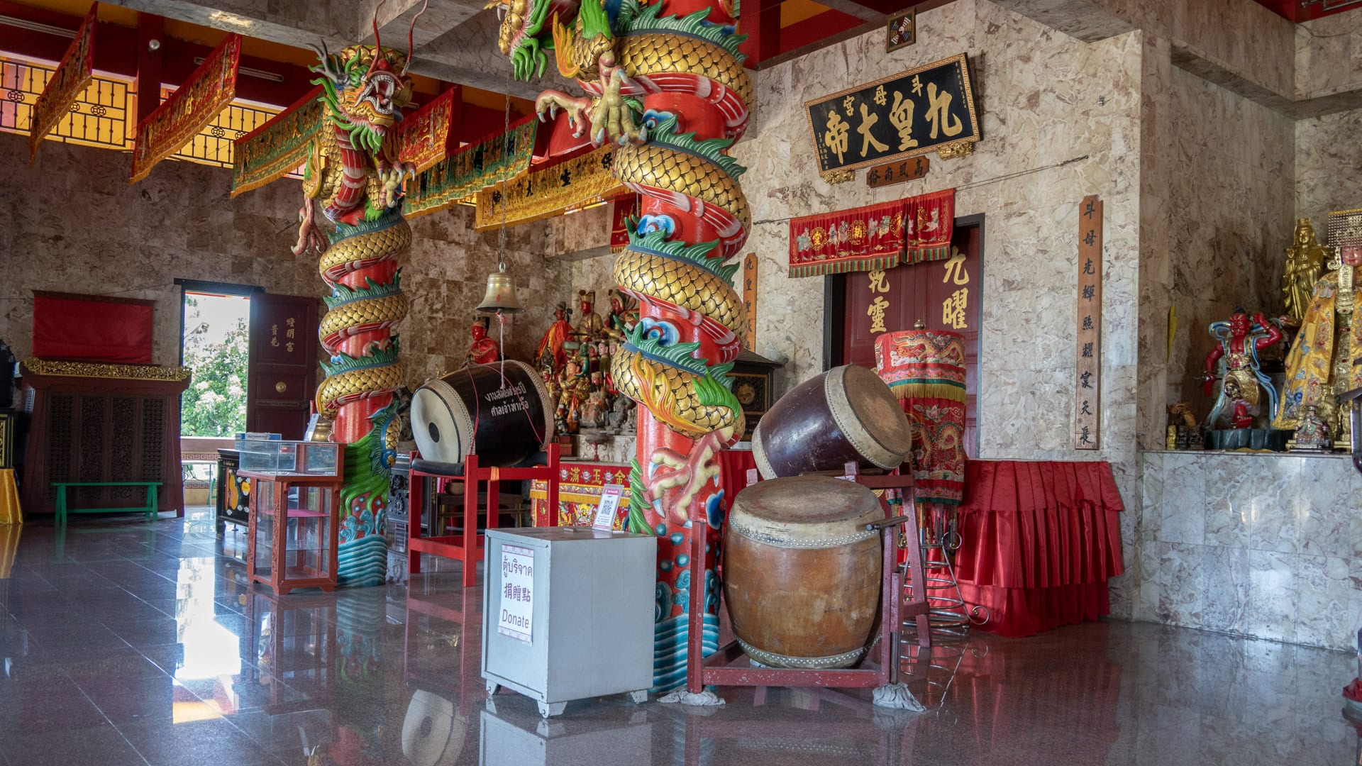 Inside San Chao Tha Ruea, two red columns wrapped with gold dragons stand beside large drums, banners, and marble-patterned walls.