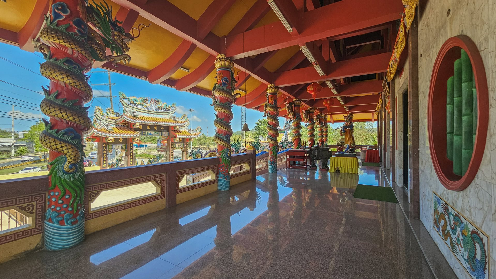 An upper veranda at San Chao Tha Ruea has dragon-wrapped columns, polished flooring, and a view out toward the entrance gate and road.