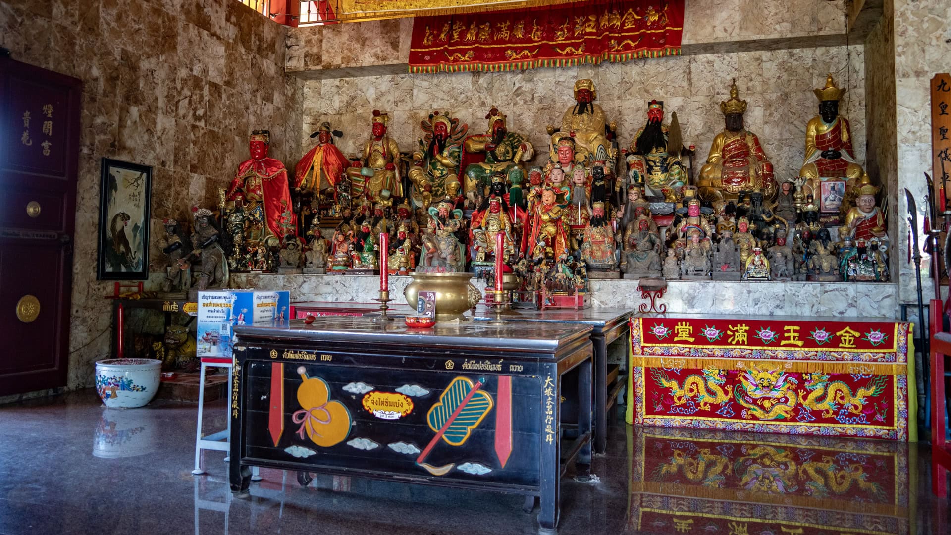 A main altar inside San Chao Tha Ruea is crowded with deity figures behind an offering table with candles and a brass incense burner.