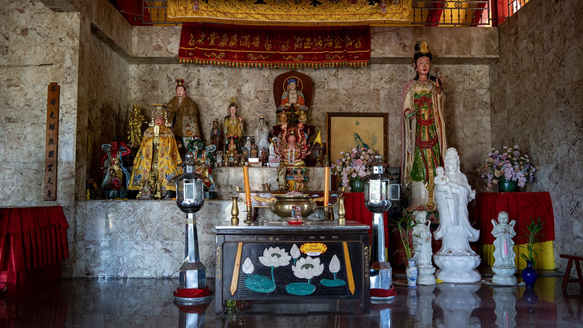 A side altar at San Chao Tha Ruea includes a tall standing Guanyin figure, smaller seated statues, lanterns, and a low offering table.