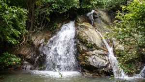 Wide waterfall cascade over tiered granite rock face into shallow green pool, dense rainforest on both sides, Kamala Phuket