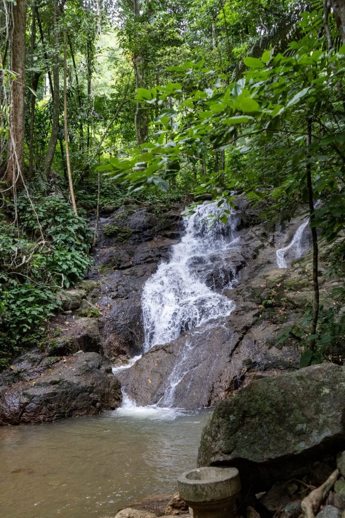 Multi-tier waterfall over dark granite into shallow pool, dense rainforest on all sides, large boulder in foreground