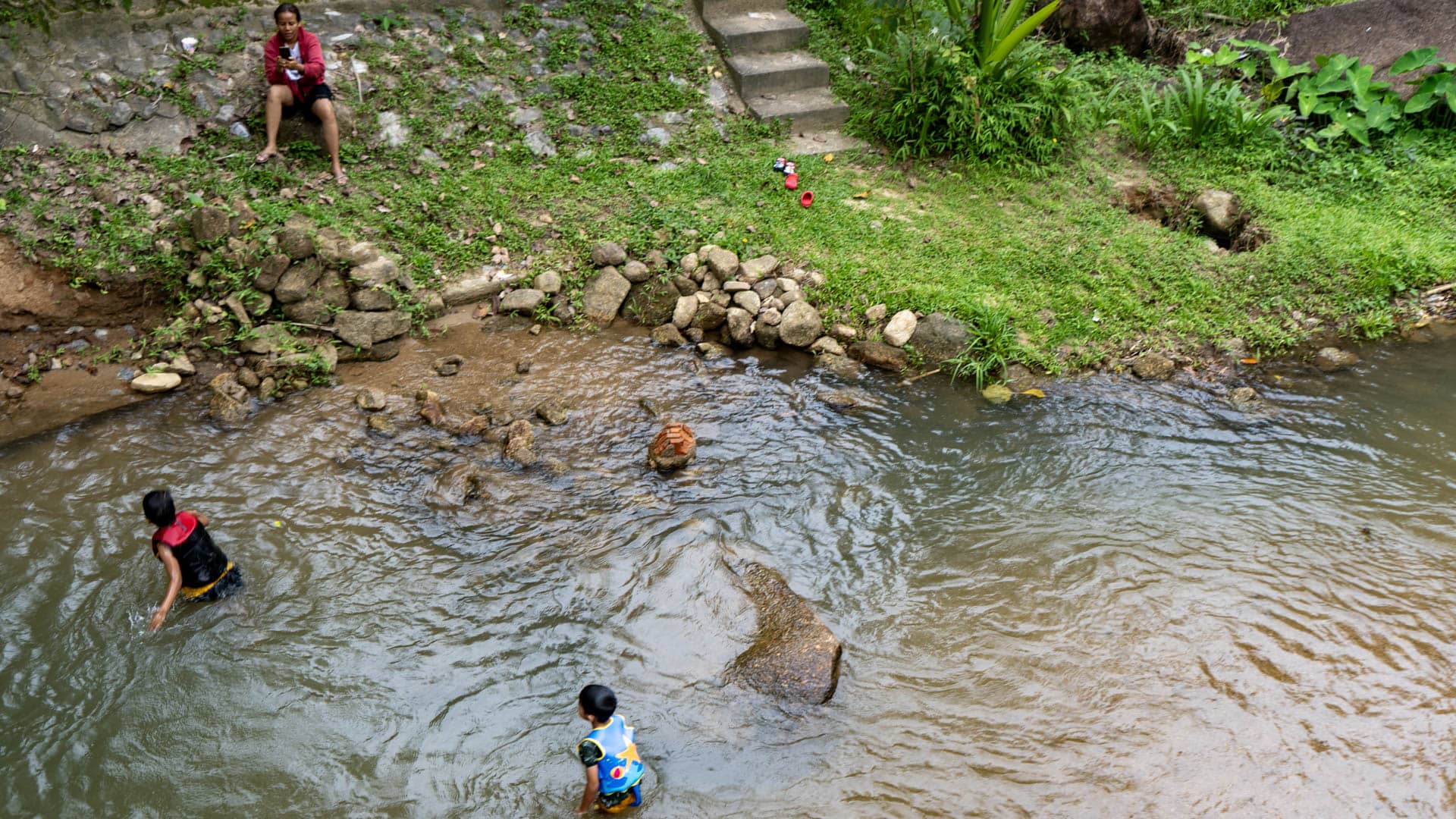 Two children wading in shallow brown-water pool, adult seated on stone bank above, tropical vegetation and concrete steps behind