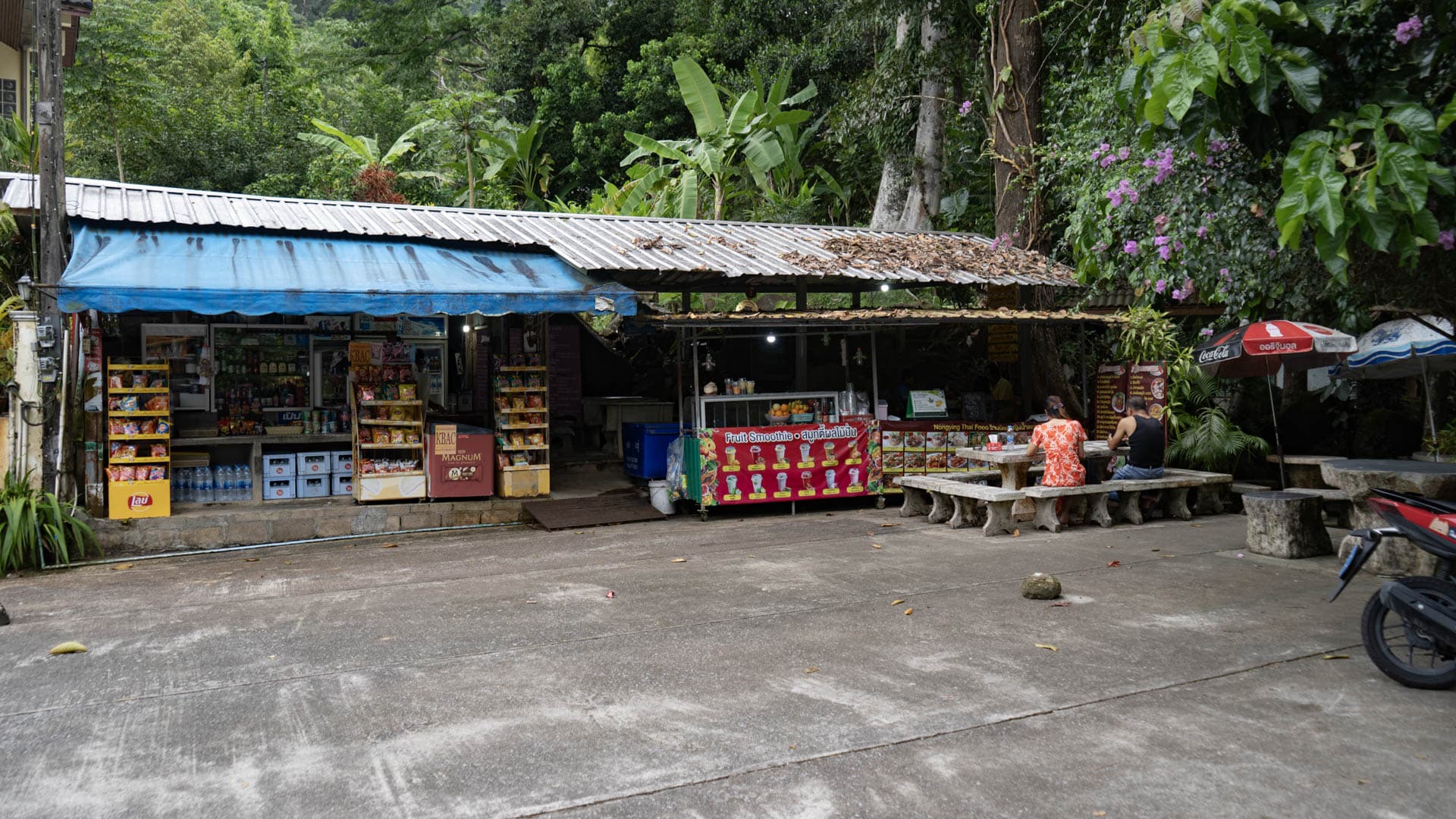Open-fronted convenience store and food stall with fruit smoothie signage, concrete forecourt, two people seated at stone bench under corrugated roof