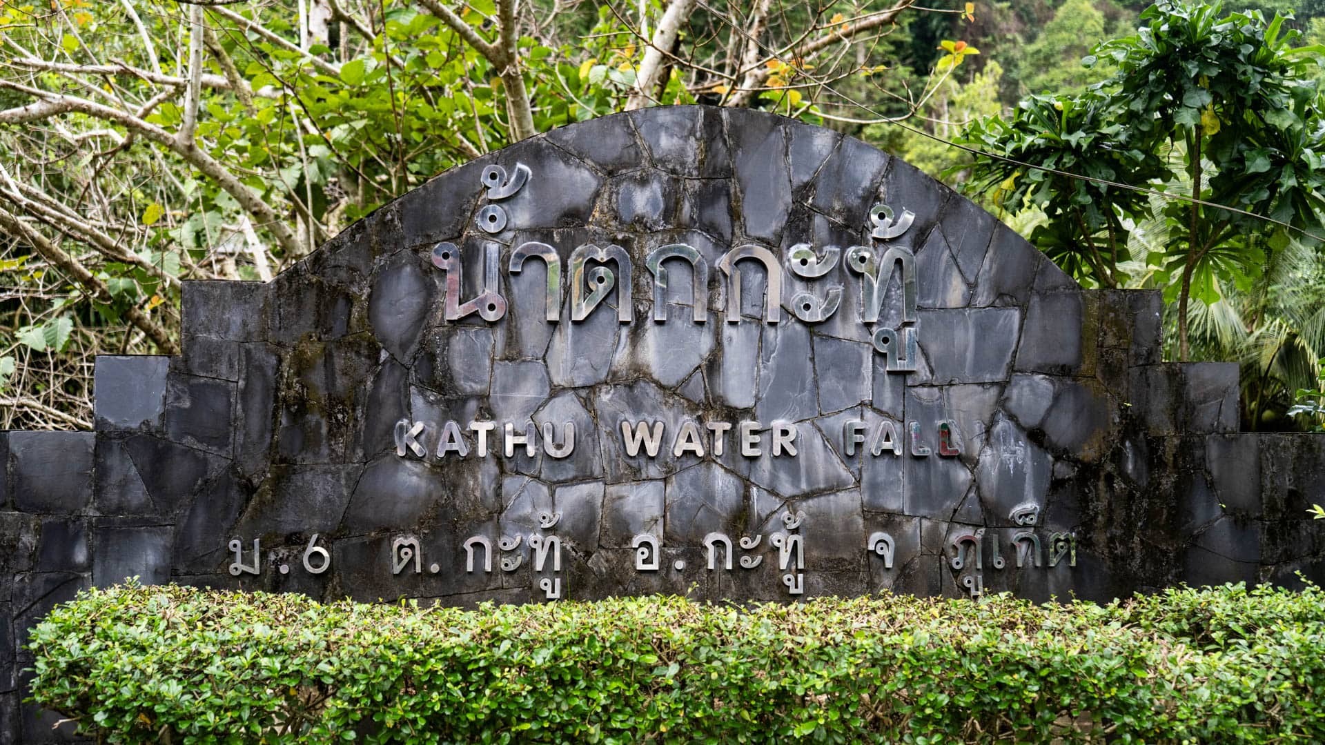Black granite arched entrance sign with Thai script above and Roman text reading Kathu Water Fall, trimmed hedge in foreground, trees behind