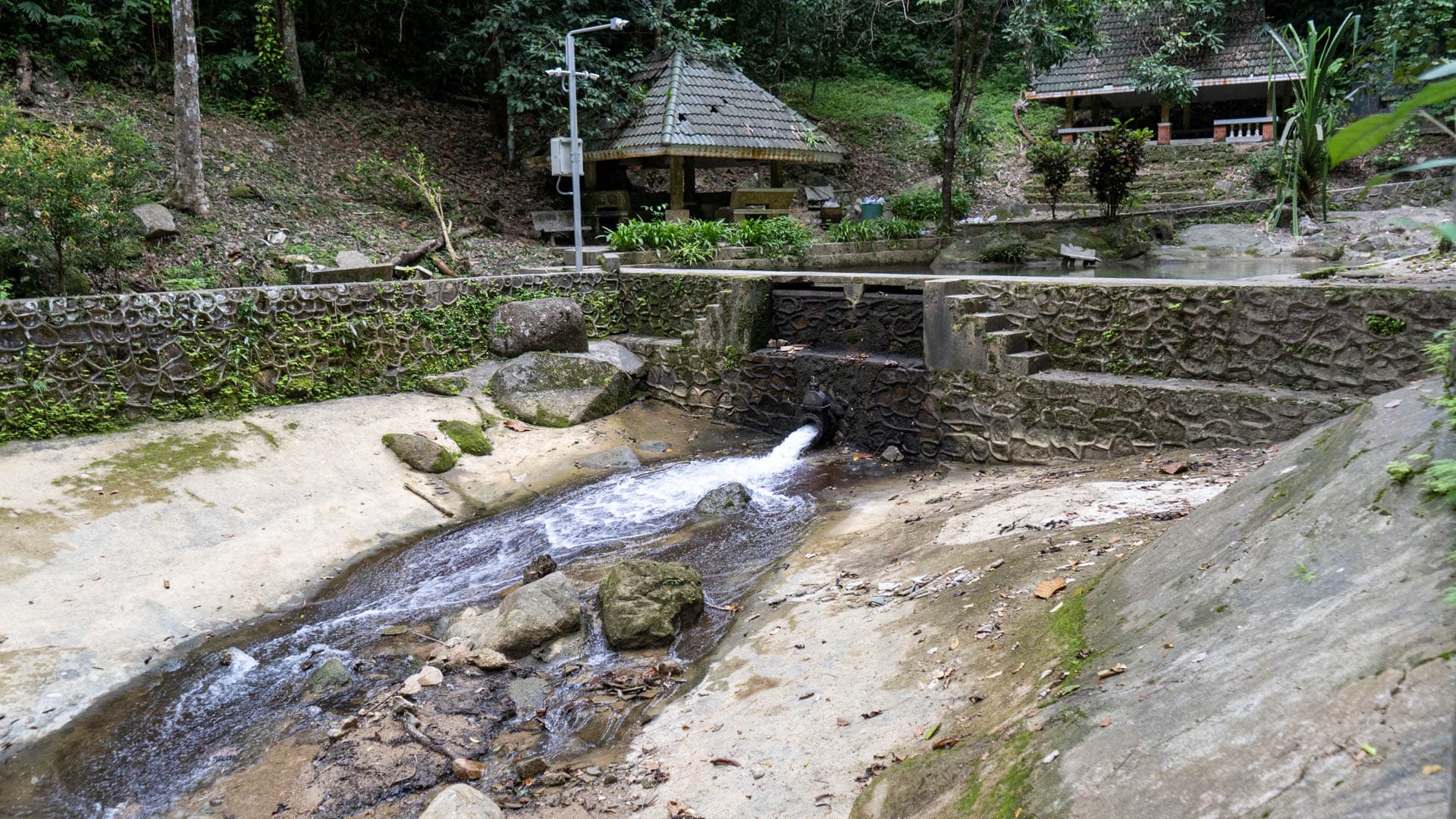 Thin stream emerging from concrete and stone weir across exposed granite rock shelf, pavilion and retaining wall in background, dry season conditions