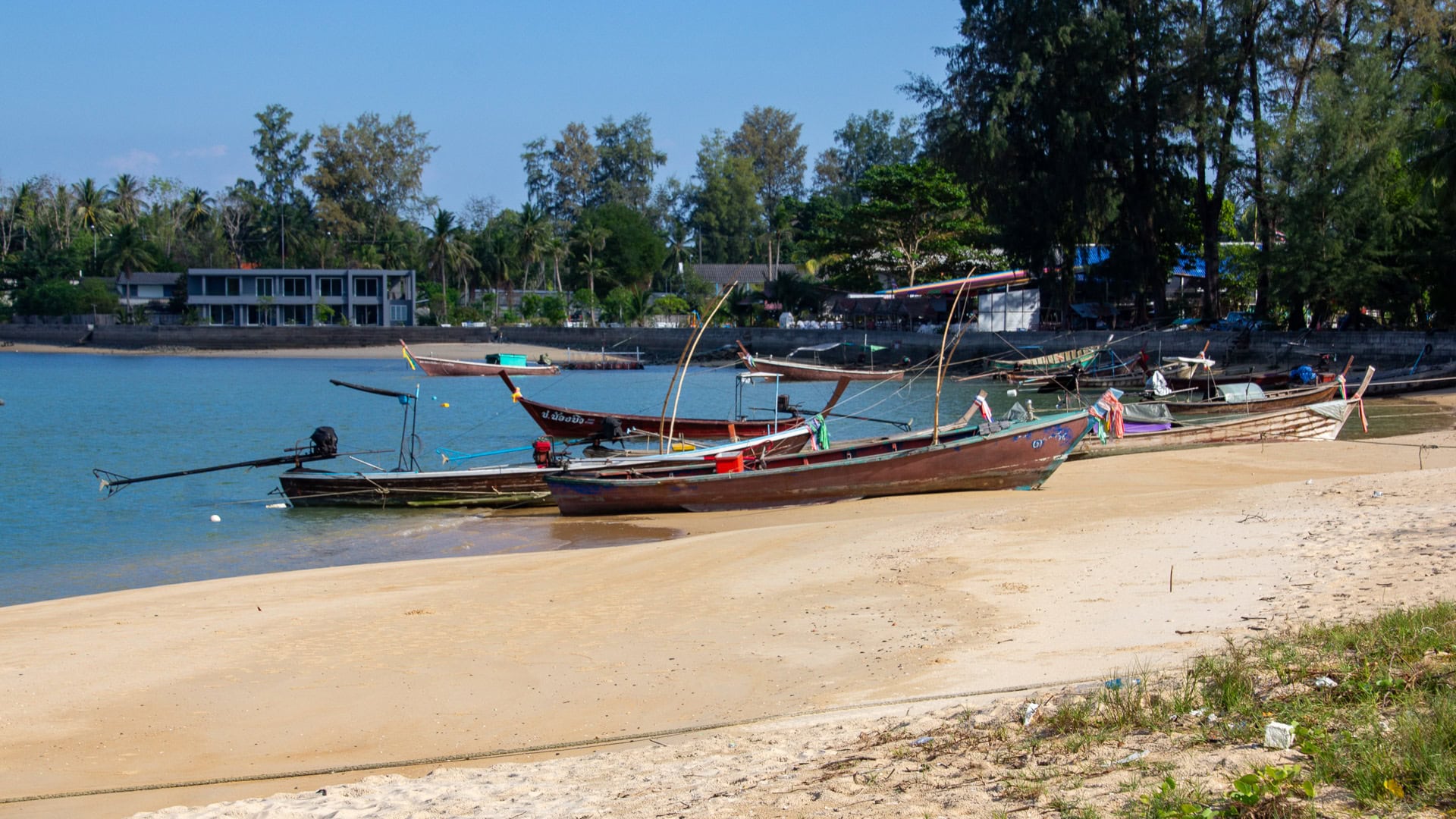 Long-tail boats resting on shallow sand at Haad Pak Phra Beach, Mai Khao, Phuket, with coastal trees in the background.