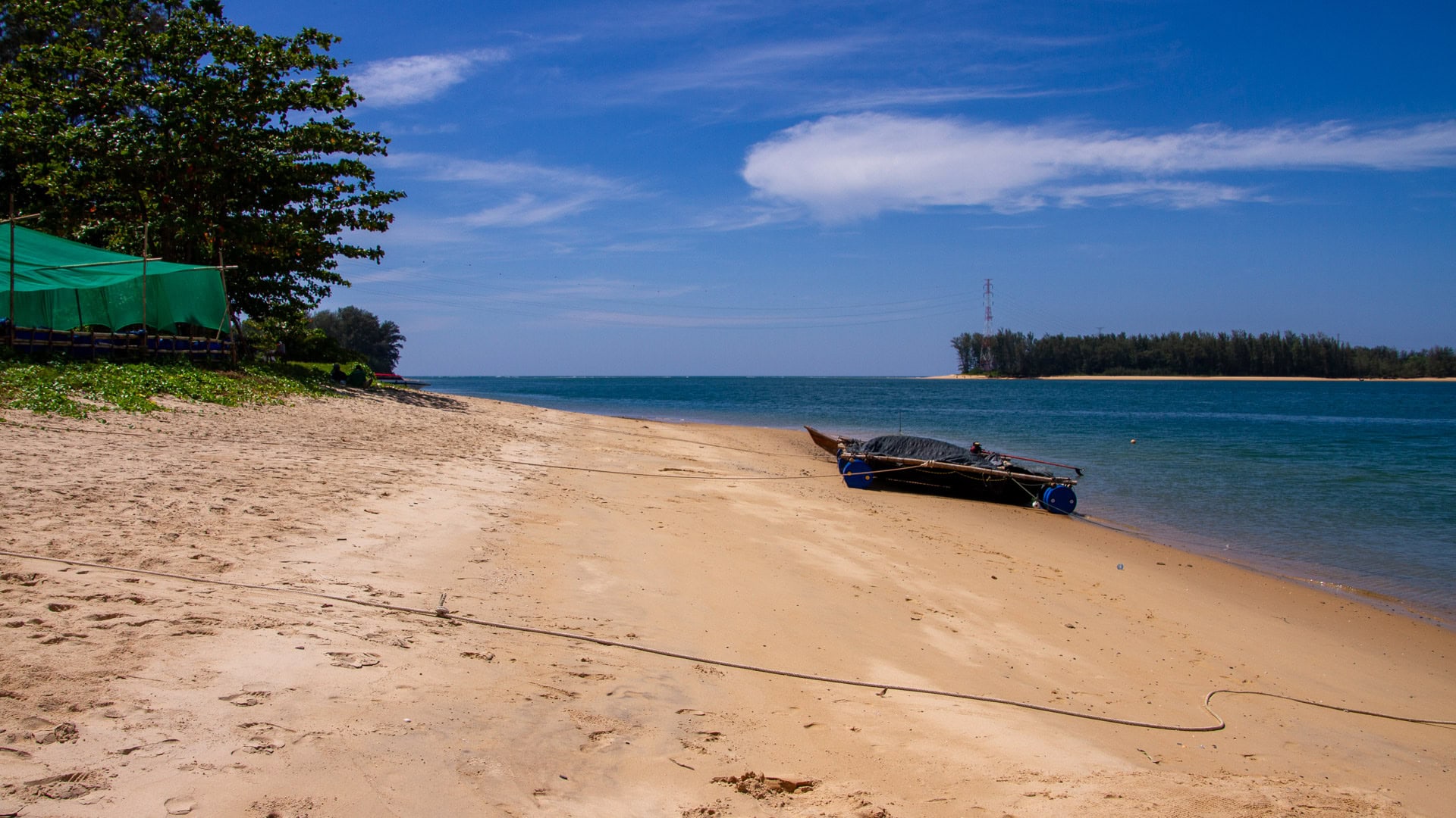 Sand shoreline at Haad Pak Phra Beach, Mai Khao, Phuket, with a long-tail boat pulled onto the beach and the strait in view.