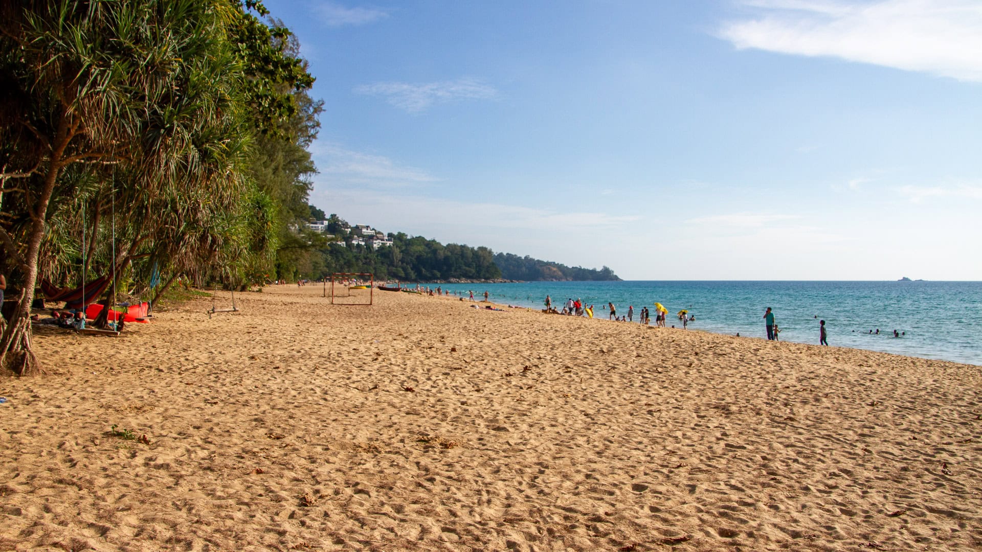 Naithon Beach Phuket long sandy shoreline with people swimming and walking, tree line on the left and open Andaman Sea on the right.
