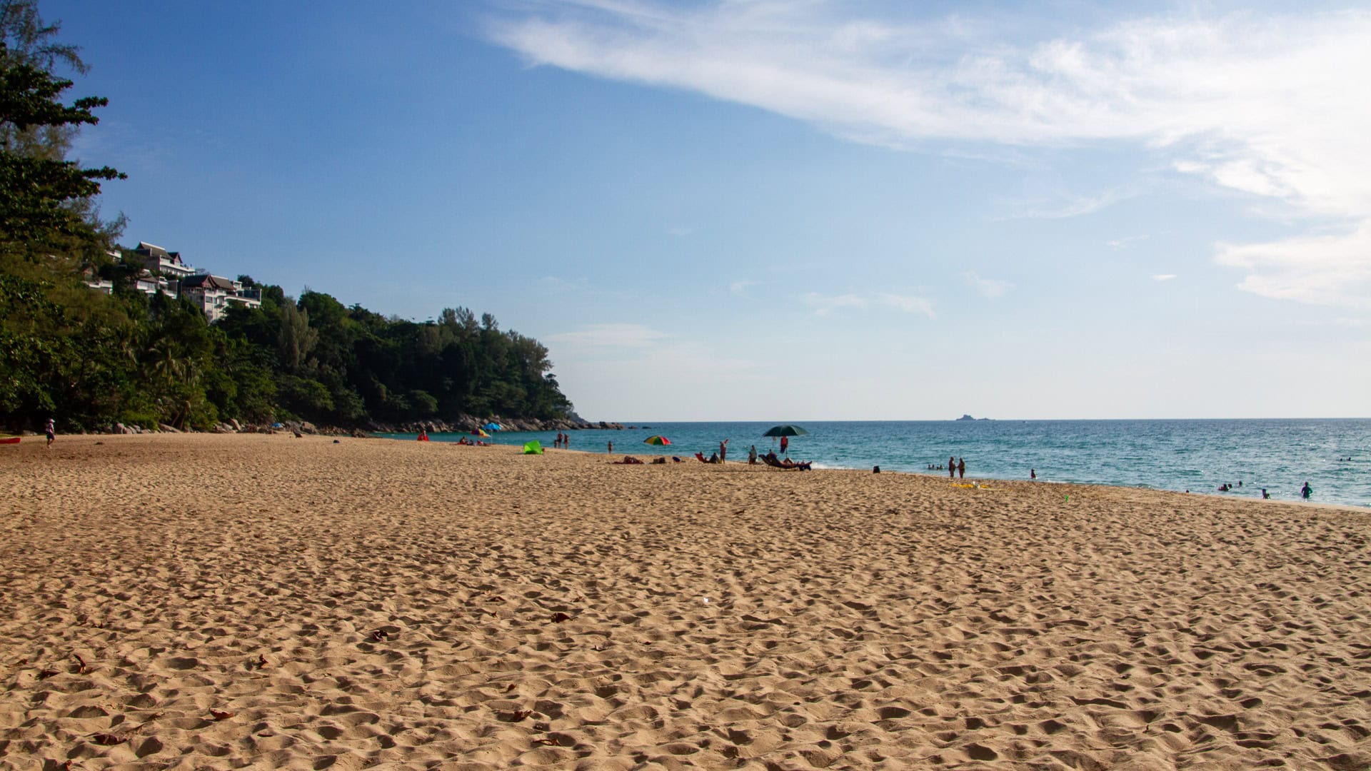 Naithon Beach Phuket broad sand and light waves with scattered swimmers and green headland closing the bay.
