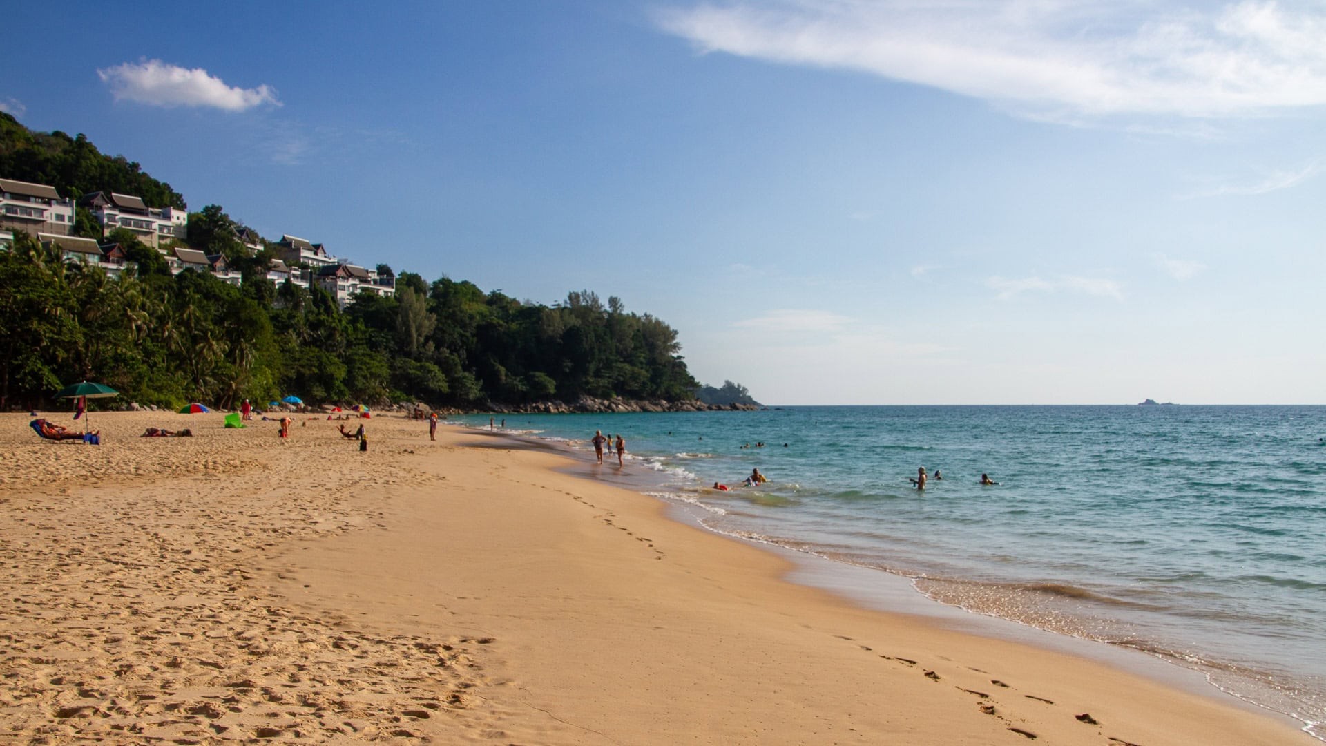 Naithon Beach Phuket view along the water’s edge with swimmers close to shore and villas perched on the forested hillside above.
