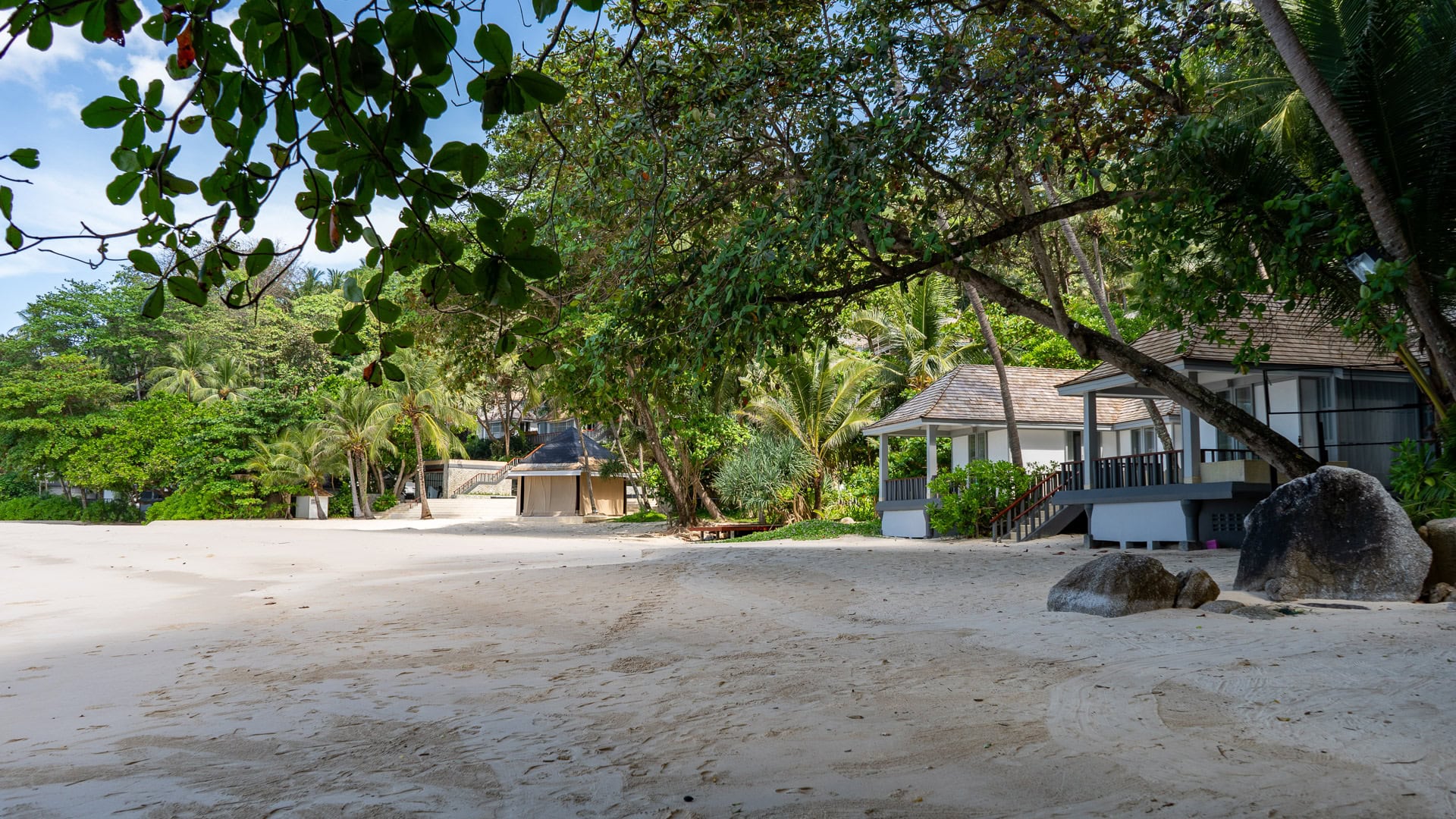 Resort villas set behind trees with shade over clean sand.